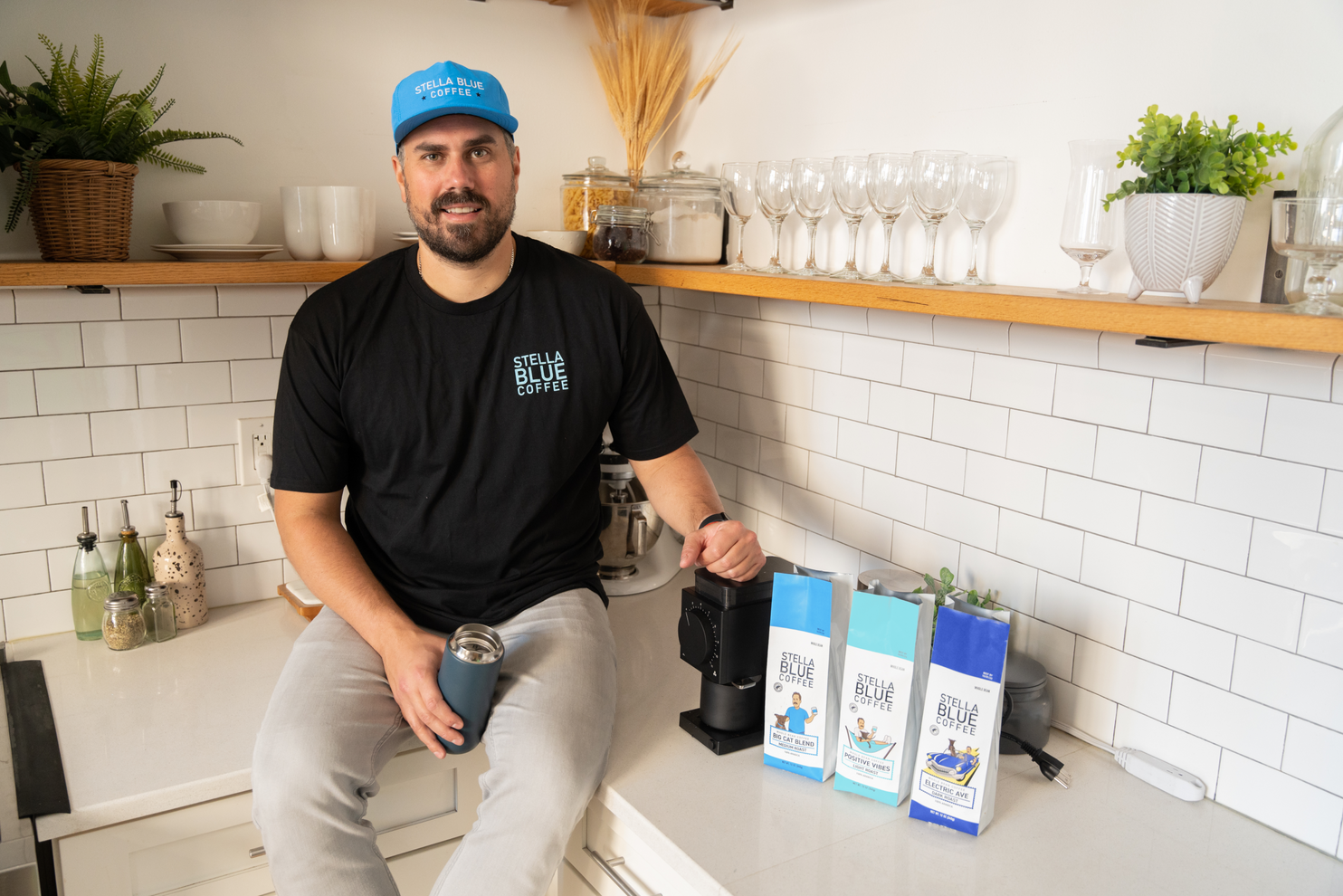 Dan Big Cat Katz sitting on a counter top holding a thermos and steadying himself against the coffee grinder with three bags of stella blue coffee